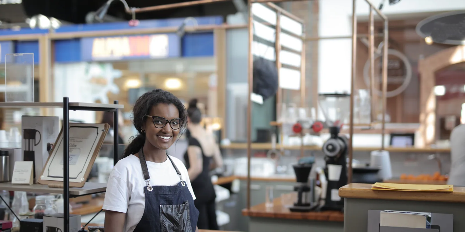 A café owner in an apron, smiling behind the counter of her shop.