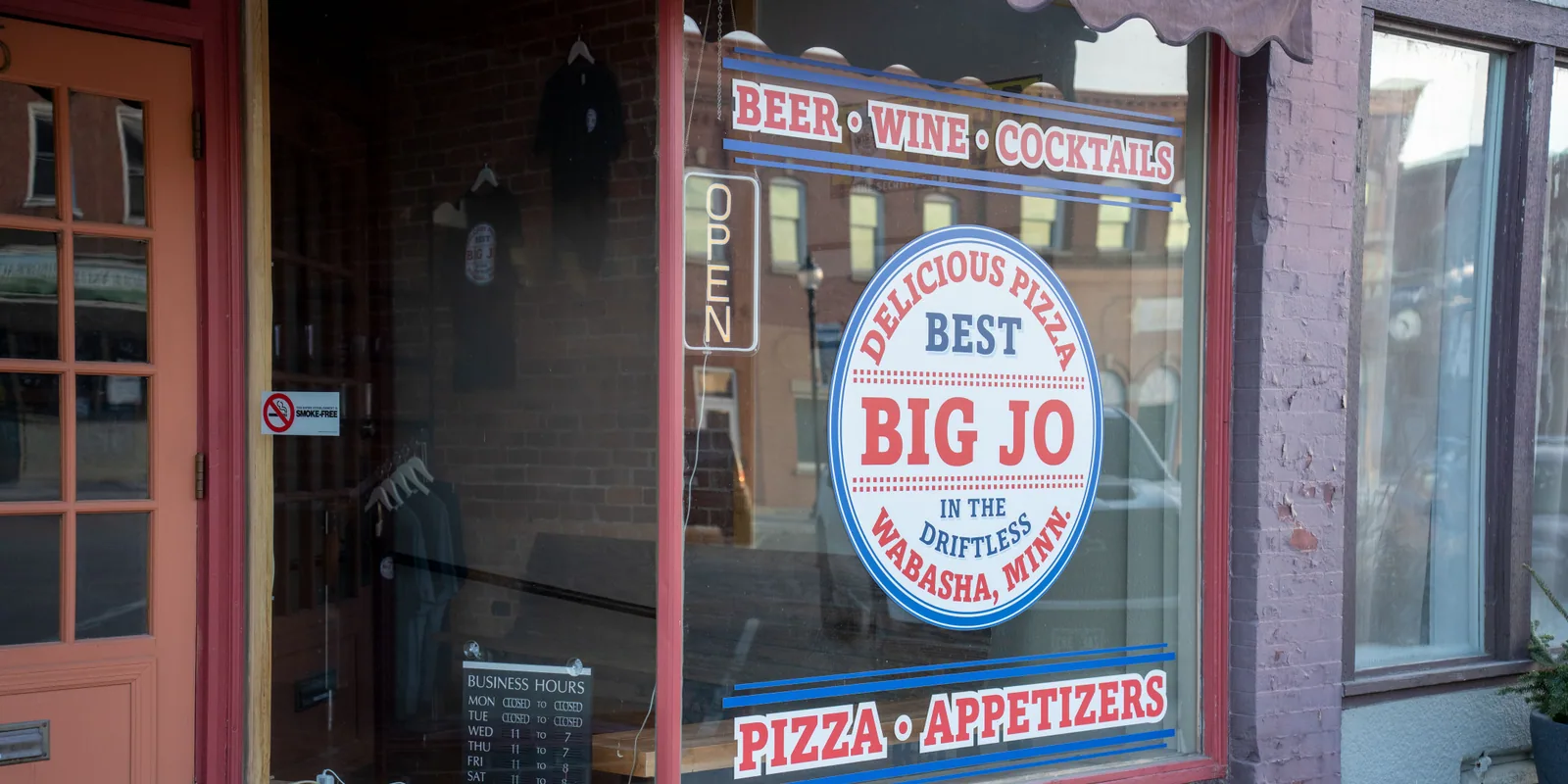 A pizza restaurant's front window with an Open sign and its business hours posted.