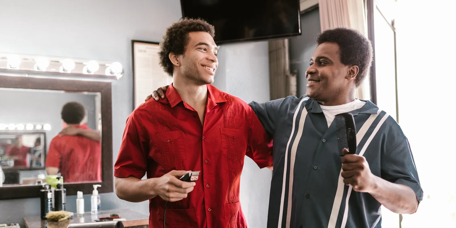 Two barbers in a shop, one with an arm around the other, both smiling.