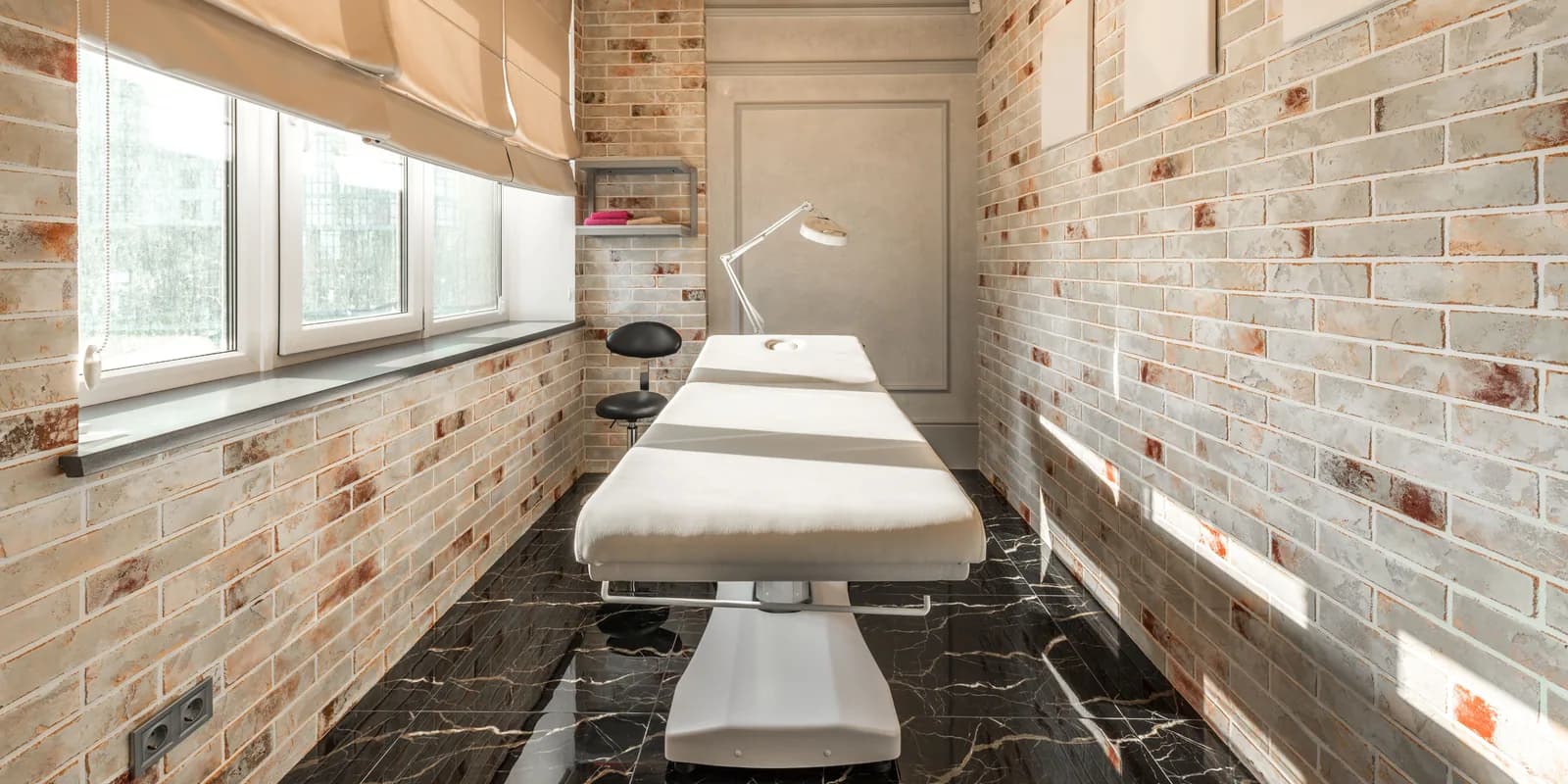 A treatment room in a modern aesthetic clinic — a bed, a lamp, brick walls and a window.