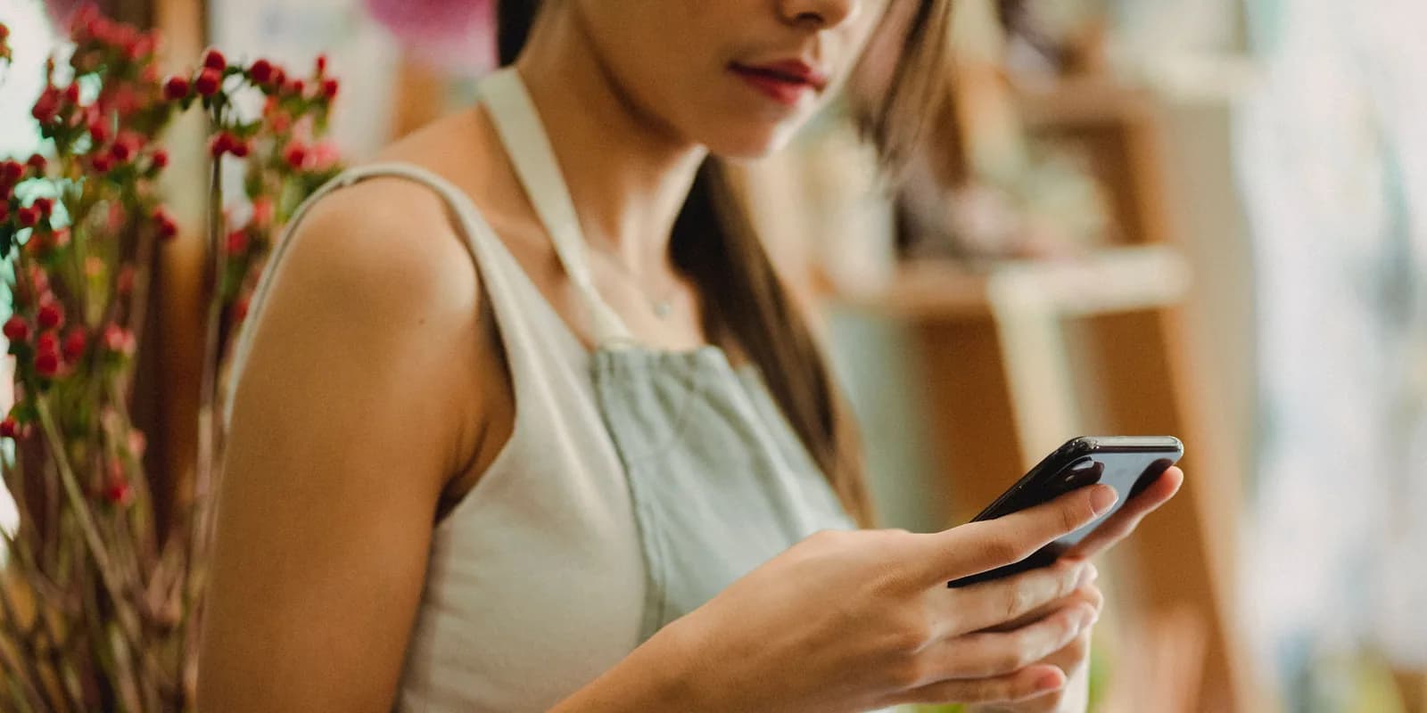 A shop owner in an apron reading her phone, flowers on the shelf behind her.